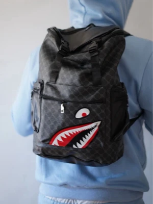 Svart ryggsäck med hajmotiv - Cool svart ryggsäck med diskret mönster och ett stort hajmotiv i rött, vitt och svart på framsidan. Ryggsäcken har flera dragkedjeförsedda fack och justerbara axelband. Perfekt för dig som vill sticka ut med en unik och trendig accessoar.