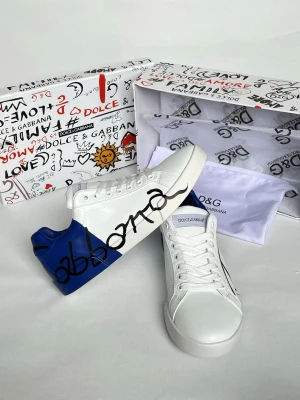 Dolce & Gabbana vita och blå sneakers - Snygga vita sneakers från Dolce & Gabbana med blå häl och cool svart grafitti-text på sidan. Skorna har klassisk låg profil, vita snören och detaljer i läder. Perfekta för dig som vill sticka ut med en unik och trendig look.