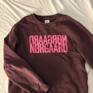 Vinröd sweatshirt från Nørgaard - Cool vinröd sweatshirt från Nørgaard med stor rosa logga framtill. Tröjan har rund halsringning, långa ärmar och ribbade muddar vid ärmslut och nederkant. Perfekt för en avslappnad och trendig look.