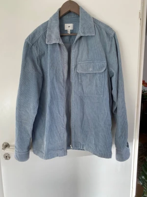 Ribbad skjorta - Tjockare blandning mellan skjorta och jacka i ljusblått ribbad tyg. Fungerar själv och som sommarjacka, men går också att para över en T-shirt eller liknande tunt plagg. Super tröja under kallare årstider.