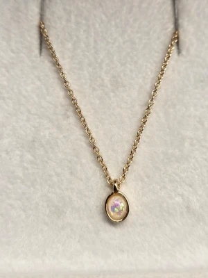 Guldhalsband med opal-liknande pärla - Snyggt halsband i guldton med en oval, skimrande opal-liknande pärla som hänge. Kedjan är tunn och elegant, och hänget har en mjuk, rundad form som fångar ljuset på ett coolt sätt. Perfekt för dig som gillar stilrena smycken med en twist. Hänget är ca 0,5 cm och kedjan 49 cm.