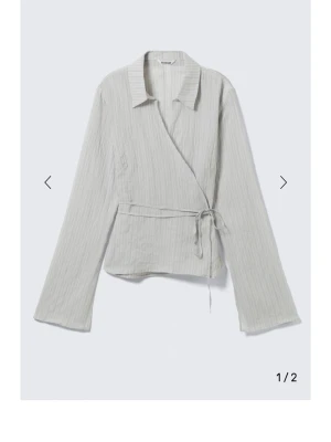 Ljusgrå omlottblus med knytband - Stilren ljusgrå omlottblus från Weekday med krage och knytband i midjan. Blusen har lång ärm.