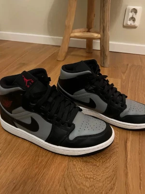 Air Jordan 1 Mid svart/grå - Snygga Air Jordan 1 Mid sneakers i svart och grått med klassisk Nike Swoosh på sidan. Skorna har svart snörning, perforerad tåbox och röd Jumpman-logga på plösen. Tillverkade i läder och syntetmaterial för en clean och sportig look.