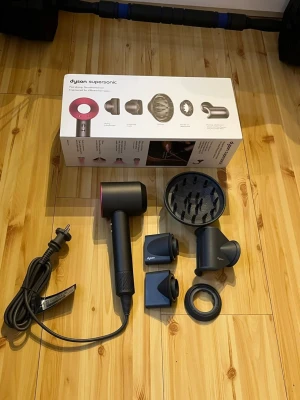 Dyson Supersonic hårfön med tillbehör - Säljer en Dyson Supersonic hårfön i svart och rosa med flera tillbehör: stylingkoncentrator, smoothing nozzle, diffuser och gentle air attachment. Modern design och kraftfull motor, perfekt för snabb och effektiv hårtorkning. Sladd och originalkartong medföljer.