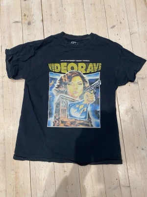 Svart Videodrome t-shirt från Rave - Cool svart t-shirt från Rave Skateboards med stort Videodrome-tryck på framsidan i retrostil. Tillverkad i 100% bomull och har klassisk passform med korta ärmar och rund hals. Perfekt för dig som gillar streetwear och grafiska prints. Står L men är M