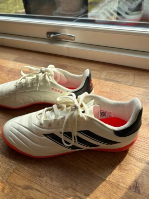 Adidas Copa vita inomhusfotbollsskor - Snygga vita Adidas Copa inomhusfotbollsskor med svarta detaljer och röda sulor. Skorna har klassisk snörning, låg profil och är tillverkade i syntetmaterial för bra komfort och grepp på inomhusplan. Perfekta för dig som vill ha stilrena och funktionella skor till futsal eller inomhusträning. Super snygga och har används fåtal gånger. De är väldigt bekväma och passar bra på foten!❣️