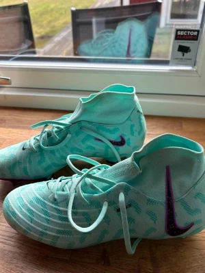 Nike Mercurial turkosa fotbollsskor - Säljer ett par Nike Mercurial fotbollsskor i en fräsch turkos färg med mönster och högt skaft. Skorna har lila Nike-logga på sidan och är tillverkade i ett lätt, syntetiskt material som ger bra bollkontroll och passform. Perfekta för dig som vill sticka ut på planen. Super snygga skor och är nästan som nya i skicket. Superbra och jätte bekväma skor!❣️