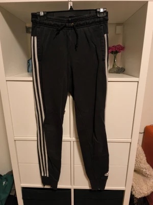 Svarta Adidas mjukisbyxor - Säljer ett par svarta mjukisbyxor från Adidas med klassiska vita ränder längs sidorna och logga vid benet. Byxorna har dragsko i midjan, muddar vid bensluten och sidofickor. Perfekta för chill eller träning.