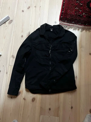 Svart overshirt från C.P. Company - Riktigt snygg overshirt/jacka passar perfekt nu när det börjar bli lite varmare och tänkte själv ha den men den passar inte längre. Den är äkta som du ser på bilden och om du är osäker på om den är äkta så scanna koden och skriv in CLG koden så ser du själv