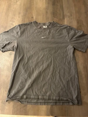 Grå Nike t-shirt med logga - En enkel grå t-shirt från Nike med den klassiska vita swoosh-loggan broderad på bröstet. T-shirten har rund halsringning och korta ärmar. Tillverkad i mjuk bomull för en skön känsla.