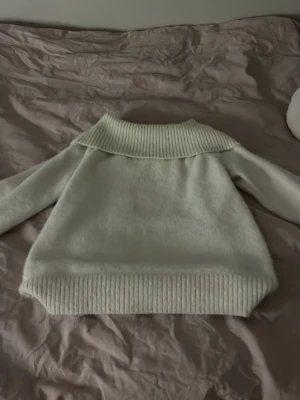 Vit stickad tröja med bred krage - Mysig vit stickad tröja med bred ribbad krage och ribbade muddar vid ärmslut och nederkant. Man kan ha den som polo men också trunt axlarna. Tröjan har en lös passform och är tillverkad i ett mjukt, fluffigt material som känns skönt mot huden. Perfekt för kyliga dagar.