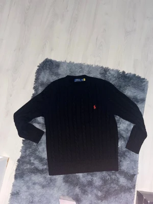 Svart kabelstickad tröja Polo Ralph Lauren - Kabelstickad svart tröja från Polo Ralph Lauren med rund halsringning och röd broderad logga på bröstet. Tröjan har ribbade muddar vid ärmslut och nederkant, perfekt för dig som gillar klassisk och stilren design.