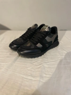 Valentino rockrunners - Säljer ett par snygga sneakers från Valentino med svart och grått camomönster. Skorna har svarta snören, detaljer i mocka och läder samt en robust sula. Perfekta för dig som gillar streetwear och vill sticka ut med exklusiva detaljer.