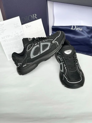 Svarta Dior sneakers med silverdetaljer - Snygga svarta Dior sneakers med silvriga detaljer och meshpaneler. Skorna har en chunky sula, svarta skosnören och tydlig Dior-logga på sidan och plösen. Perfekta för dig som vill ha en exklusiv och modern look.