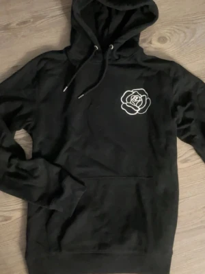Svart hoodie med ros och text - Svart hoodie med stor huva och vitt tryck av en ros. Hoodien har magficka och ribbade muddar. Perfekt för dig som gillar streetwear och grafiska tryck. Aldrig använd, från trycklagret 🌹✨ Passar mig perfekt som vanligt vis har XS, S.