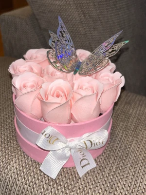 Rosa presentbox med rosor och fjäril - Söt rund presentbox i rosa med konstgjorda ljusrosa rosor och en dekorativ holografisk fjäril på toppen. Boxen är dekorerad med ett vitt band med logga. Perfekt som dekoration eller gåva.