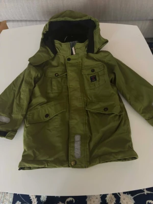 Grön parkasjacka från Polarn O. Pyret - Stilren grön parkasjacka från Polarn O. Pyret med huva, fyra framfickor med knappar och reflexdetaljer på ärm och framtill. Jackan har en rak passform och är perfekt för kyliga dagar. Materialet är slitstarkt och vattenavvisande.