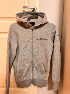 Grå hoodie Peak Performance - Snygg grå hoodie från Peak Performance med dragkedja framtill och två fickor. Märkets logga i svart på bröstet och en liten logga på ärmen. Mjuk bomullsblandning, ribbade muddar och huva för extra komfort.