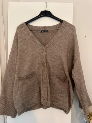 Beige kofta från SHEIN med fickor - Mysig beige kofta från SHEIN med v-ringning, knäppning framtill och två stora fickor. Koftan har breda ribbade muddar vid ärmslut och nederkant samt en avslappnad passform. Perfekt för lager på lager och chill dagar.