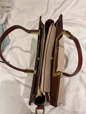 Beige och brun handväska med gulddetaljer - Snygg handväska i beige och brunt med guldfärgade metalldetaljer och två rejäla handtag. Väskan har flera fack, inklusive ett innerfack med dragkedja. Materialet ser ut att vara konstläder med struktur på utsidan. Perfekt för dig som gillar stilrena accessoarer.