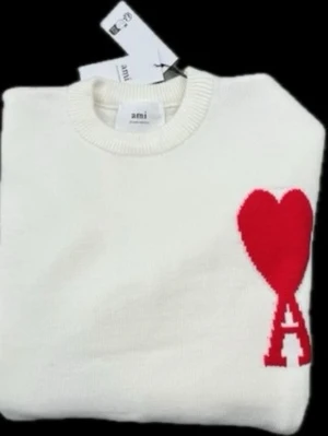 Vit Ami Paris - Säljer en vit stickad tröja från Ami Paris med rund halsringning och ribbade muddar. Tröjan har ett stort rött hjärta med bokstaven 'A' broderat på bröstet, vilket ger en unik och trendig look. Perfekt för dig som gillar stilrena plagg med en twist.