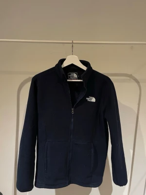 Marinblå fleecejacka The North Face - Mysig marinblå fleecejacka från The North Face Summit Series med dragkedja framtill och två fickor. Jackan har hög krage och broderad logga på bröstet. Perfekt för lager-på-lager och outdoor-looks.