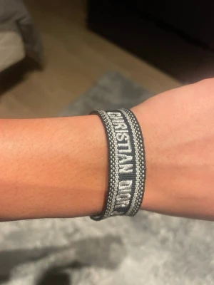  Christian Dior tygarmband - Snyggt grått tygarmband från Christian Dior med texten 'Christian Dior J'Adior' broderad i vitt. Armbandet har en stilren och modern design med kontrasterande kanter och justerbar knytning. Perfekt accessoar för att lyfta din outfit.