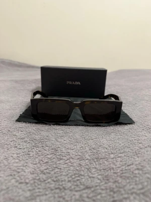 Prada rektangulära solglasögon brun - Snygga solglasögon från Prada med rektangulär form och mörkbruna bågar i ett sköldpaddsmönster. Glasen är mörka och bågarna har Prada-loggan på sidan. Perfekta för dig som vill ha en trendig accessoar med lyxig känsla.