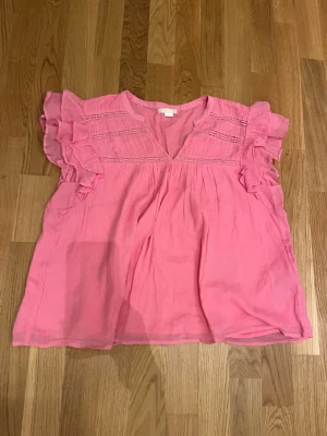 Rosa blus Gina Tricot - Söt rosa blus med v-ringning och volangdetaljer på ärmarna. Blusen har ett luftigt och flowy material, samt dekorativa sömmar upptill. Perfekt för dig som gillar en feminin och avslappnad stil.