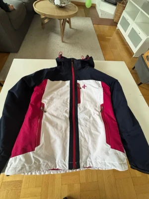 Cross Sportswear jacka  - Jacka från Cross Sportswear i marinblått, vitt och ceriserosa. Utrustad med huva, dragkedja framtill och två stora fickor med dragkedja. Sportig design med logga på bröstet och detaljer i syntetmaterial. 