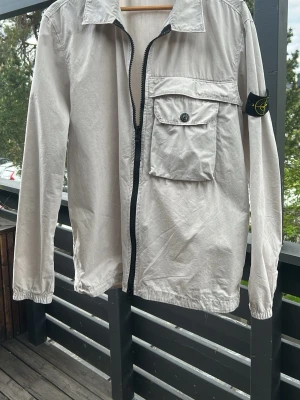 Beige overshirt från Stone Island - Snygg beige overshirt från Stone Island med klassisk svart och gul logotyp på ärmen. Jackan har dragkedja framtill, en stor bröstficka med knapp och elastiska muddar vid ärmslut. Jackan är något mindre i storleken. Hör av er om ni har frågor! 