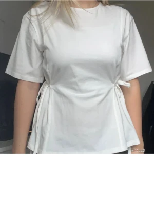 Vit t-shirt med knyt i sidan - Snygg vit t-shirt med korta ärmar och knyt i sidorna. Köpt på plick och två första bilderna är lånade, passade tyvärr inte mig och säljer därför vidare. Köpte för samma pris som jag säljer den för. Inga defekter