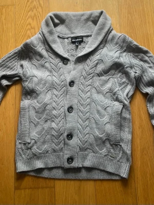 Grå kabelstickad kofta från Threadbare - Snygg grå kabelstickad kofta från Threadbare med bred krage och knappar framtill. Koftan har två fickor och ribbade muddar vid ärmslut och nederkant. Perfekt för lager-på-lager och en chill look.