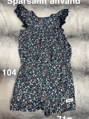 Blommig jumpsuit från Newbie - Supersöt blommig jumpsuit från Newbie i mörkblått med små blommor i pastellfärger. Volangärmar och resår i midjan ger en fin passform. Perfekt för varma dagar och enkel att matcha med sneakers eller sandaler.🌸 Sparsamt använd. Storlek 104 