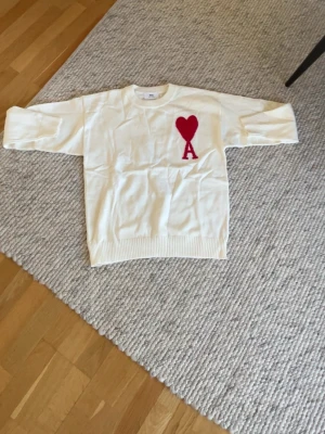 Vit stickad tröja från Ami Paris - Vit stickad tröja från Ami Paris med rött hjärta och bokstaven A på bröstet. Tröjan har rund halsringning, ribbade muddar och är gjord i mjukt material. Perfekt för dig som gillar stilrena och trendiga plagg.