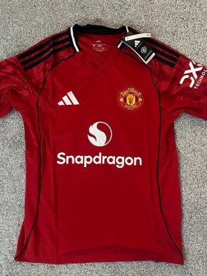 Manchester United matchtröja Adidas M - Röd Manchester United matchtröja från Adidas i slim fit-modell. Tröjan har klubbmärke på bröstet, svarta och vita detaljer på axlarna samt sponsorloggor framtill och på ärmen. Tillverkad i lätt och ventilerande material, perfekt för fotbollsträning. Bara skicka meddelande vid frågor 👍