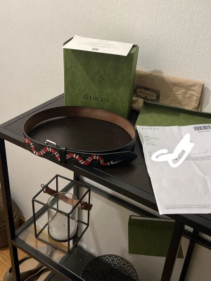 Gucci svart bälte med röd/grön detalj - Snyggt svart bälte från Gucci med klassiska röd- och grönrandiga detaljer. Bältet har en silverfärgad metallspänne med Gucci-logga och är tillverkat i läder. Perfekt accessoar för att lyfta din outfit. Box och kvitto medföljer 