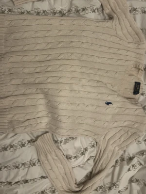 Vit kabelstickad tröja Polo Ralph Lauren - Kabelstickad vit tröja från Polo Ralph Lauren med rund halsringning och blå broderad logga på bröstet. Tröjan har långa ärmar och ribbade muddar vid ärmslut och nederkant. Klassisk och tidlös design i mjukt material.