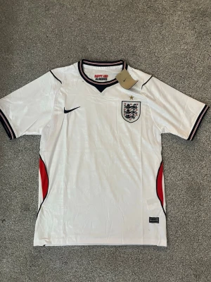 England Nike fotbollströja vit VM2026(Herr) - Vit England fotbollströja från Nike med broderat landslagsmärke och svart Nike-logga på bröstet. Tröjan har svarta, röda och vita detaljer vid krage och ärmslut samt röda inslag vid sidorna. Tillverkad i Dri-FIT material för bra ventilation. Bara skicka meddelande vid frågor👍