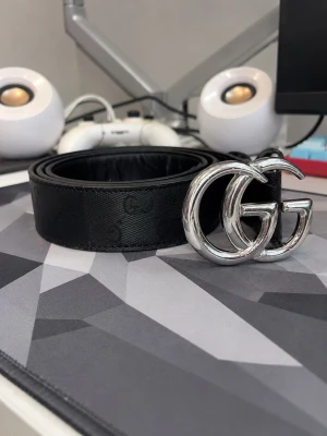 Svart  bälte med silver-spänne - Snyggt svart bälte från med klassisktmönster och ett stort, glansigt silverfärgat -spänne. Bältet är gjort i skinn och har en stilren look som passar till många outfits. Man ser på bilden att de finns ett hål som är lite trasigt,men det är inget som syns utifrån eller påverkar bältet 