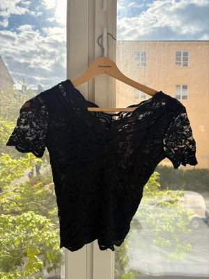 Svart spetsblus med v-ringning - Snygg svart blus i spets med v-ringning och korta ärmar. Blusen har ett feminint blommigt spetsmönster och är lätt transparent. Perfekt att styla med jeans eller kjol för en trendig look.