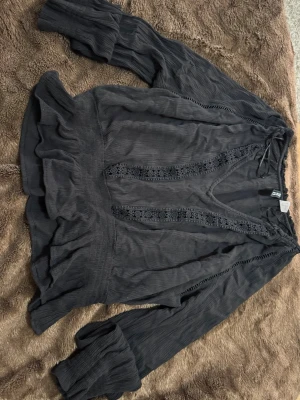 Svart blus med spetsdetaljer H&M - Svart blus från H&M Divided med långa ärmar och v-ringning. Blusen har fina spetsdetaljer längs framsidan och volang vid nederkanten. Tillverkad i ett lätt, strukturerat tyg som ger en avslappnad men ändå stilren look.