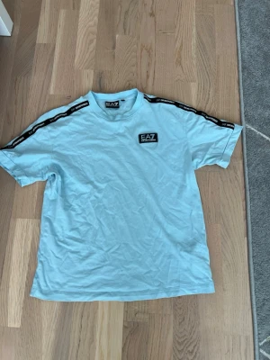 Ljusblå EA7 Emporio Armani t-shirt - Snygg ljusblå t-shirt från EA7 Emporio Armani med logga på bröstet och svarta band med vit text längs axlarna. Klassisk passform och rund hals. Perfekt för en trendig och sportig look.