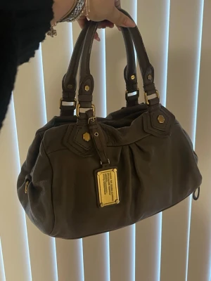 Marc Jacobs handväska - Snygg brun/beige handväska i mjukt läder från Marc by Marc Jacobs. Väskan guldfärgade metalldetaljer och en ikonisk metallbricka framtill. Perfekt för dig som vill ha en stilren och klassisk accessoar. Pris kan diskuteras vid snabb affär ⚡️👜💰