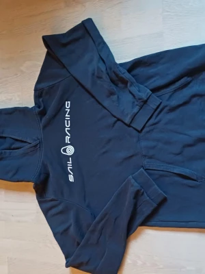 Mörkblå Sail Racing hoodie med tryck - Cool mörkblå hoodie från Sail Racing med vitt logotryck på bröstet. Klassisk huva och känguruficka framtill. Mjukt material och ribbade muddar vid ärmslut och nederkant. Perfekt för dig som gillar sportig stil.
