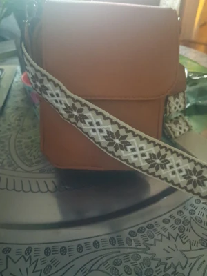 Brun axelväska med mönstrat band - Snygg brun axelväska i slät konstläder med lock och bred axelrem i beige och brunt med coolt mönster. Perfekt för dig som vill ha en stilren men ändå unik väska till din outfit.