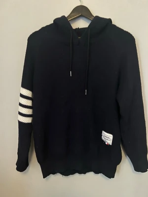 Svart hoodie från Thom Browne - Svart hoodie från Thom Browne med klassiska vita ränder på ena ärmen och dragsko i huvan. Stickad känsla och liten etikett nertill med varumärket. Perfekt för en stilren och trendig look.
