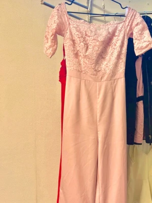Ljusrosa off shoulder jumpsuit spets - Supersöt ljusrosa jumpsuit med off shoulder-modell och korta ärmar. Överdelen är i blommig spets och byxorna är vida och släta. Perfekt för dig som vill sticka ut med en romantisk look. Dragkedja bak för enkel påklädning.