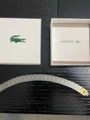 Silverfärgat armband från Lacoste - Stilrent armband från Lacoste i silverfärgad metall med rektangulära länkar och diskret Lacoste-logga. Perfekt för dig som gillar minimalistisk design och vill ha ett snyggt accessoar till din outfit. Levereras i originalask med Lacoste-märke.