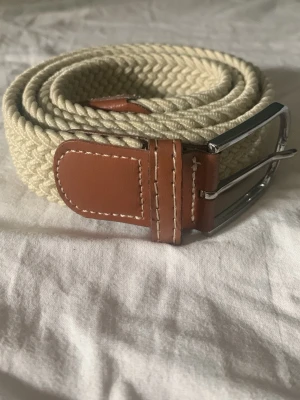 Beige flätat bälte 85 cm - Snyggt beige flätat bälte med bruna skinn-detaljer och silverfärgat spänne. Perfekt accessoar för att lyfta din outfit med en stilren och klassisk look. Bältet har en flexibel design som gör det enkelt att justera.